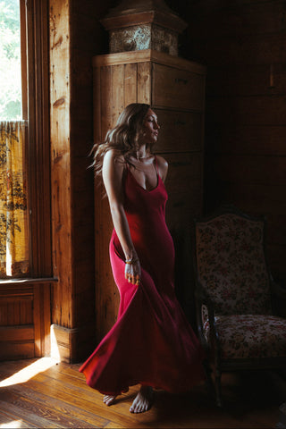 Woman in a red dress standing in a sunlit room with wooden walls and a window.