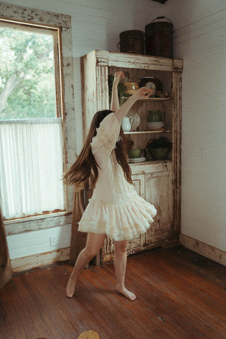 Woman in a white dress standing in a room with wooden floors and a large window.