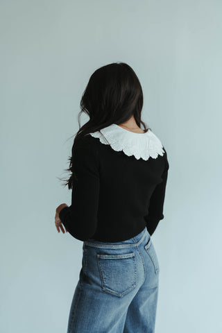Person wearing a black sweater with a white collar and blue jeans on a light gray background