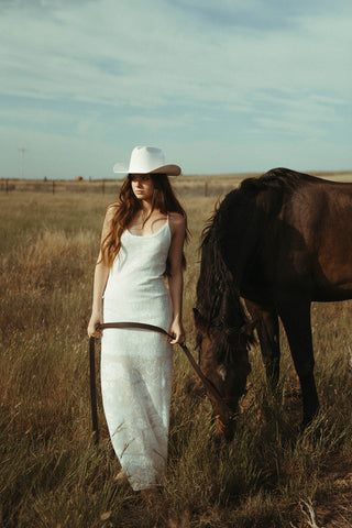 Woman in a white dress and cowboy hat standing next to a horse in a field.