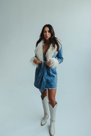 Woman wearing a denim dress with fur trim and white cowboy boots on a light gray background