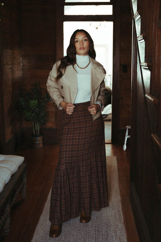 Woman standing in a wooden interior space with a plaid skirt and beige jacket.