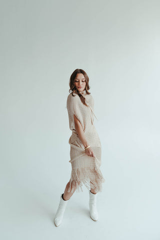Woman in a beige dress with fringe and white boots standing against a plain background