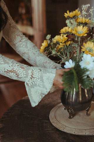 Person wearing a lace sleeve holding a bouquet of flowers with a vase on a table.