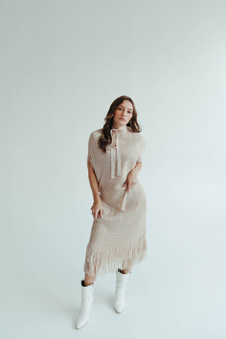 Woman wearing a beige knit dress with fringe details and white boots on a light background
