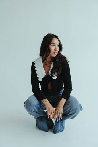 Woman wearing a black cardigan with white trim and blue jeans on a light gray background