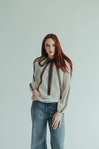 Woman wearing a white blouse with black details and blue jeans against a light gray background