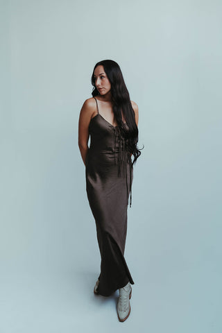Woman wearing a long brown dress with lace-up details against a light gray background